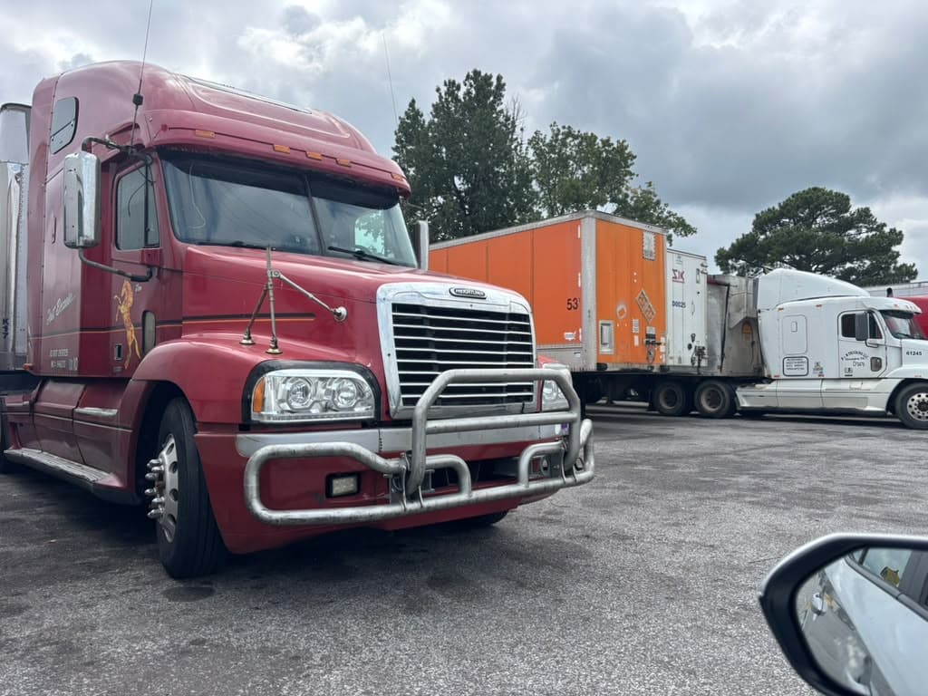 Red commercial highway tractor equipped with front deer guard parked beside freight trailers transporting regulated cargo